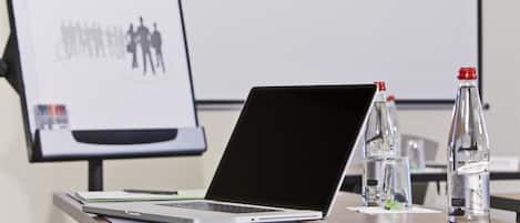 Ein Laptop auf einem Schreibtisch, im Hintergrund ein Präsentationsbildschirm, Wasserflaschen und ein Notizbuch.