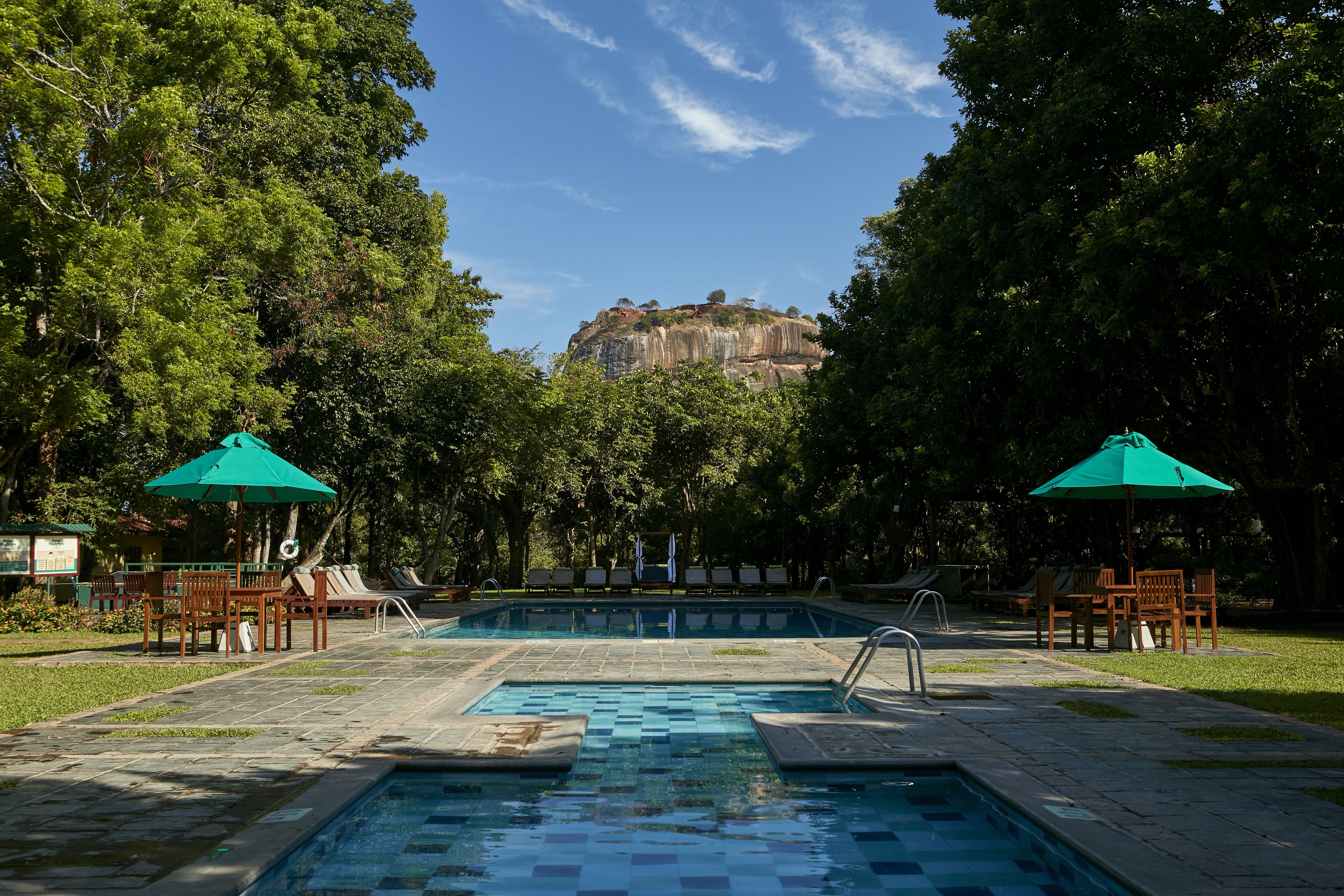 Piscina con sillones de descanso y sombrillas, rodeada de árboles y con vista a una montaña.