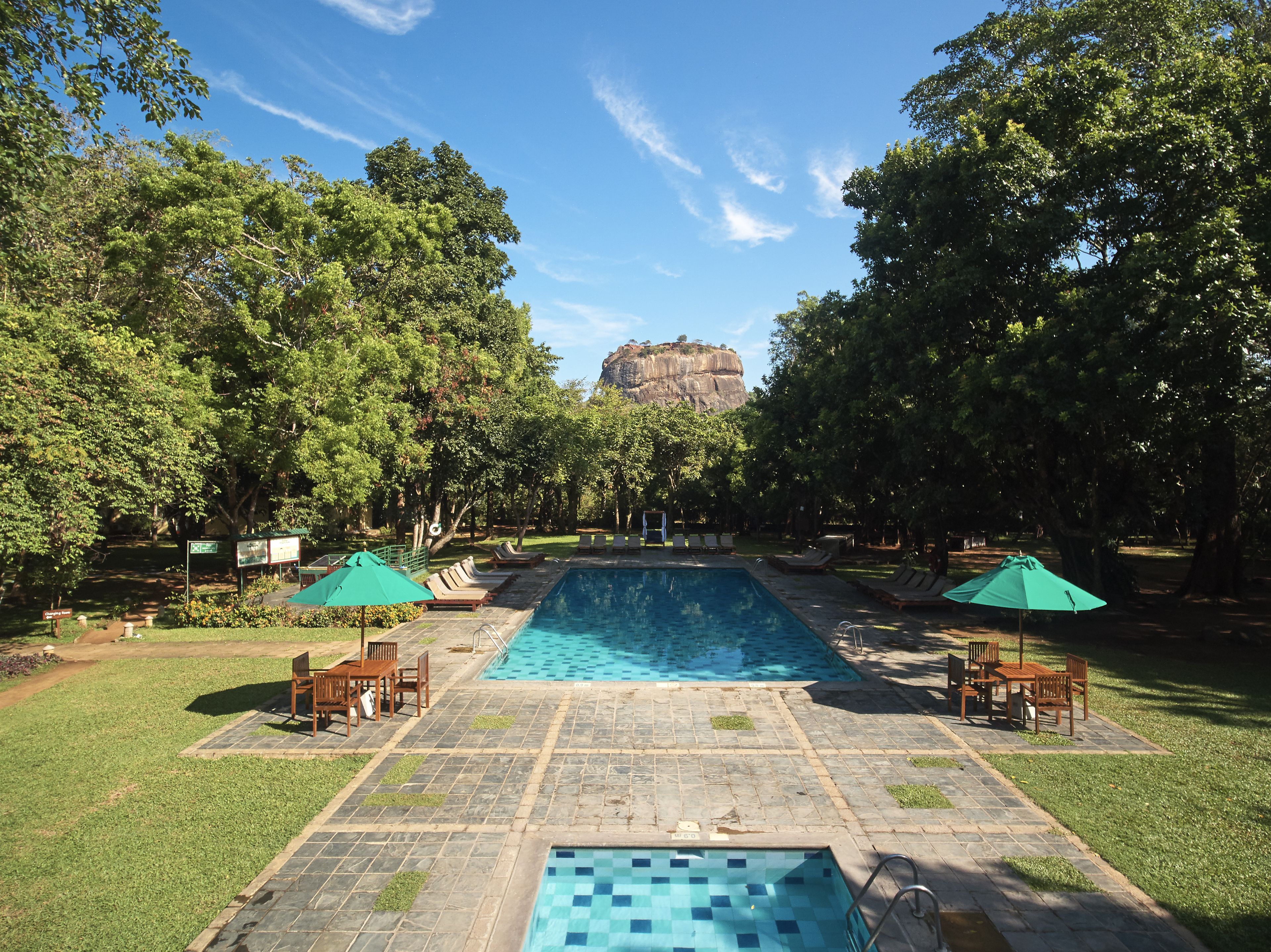 A swimming pool with lounge chairs and umbrellas, surrounded by trees and a rocky cliff in the background.