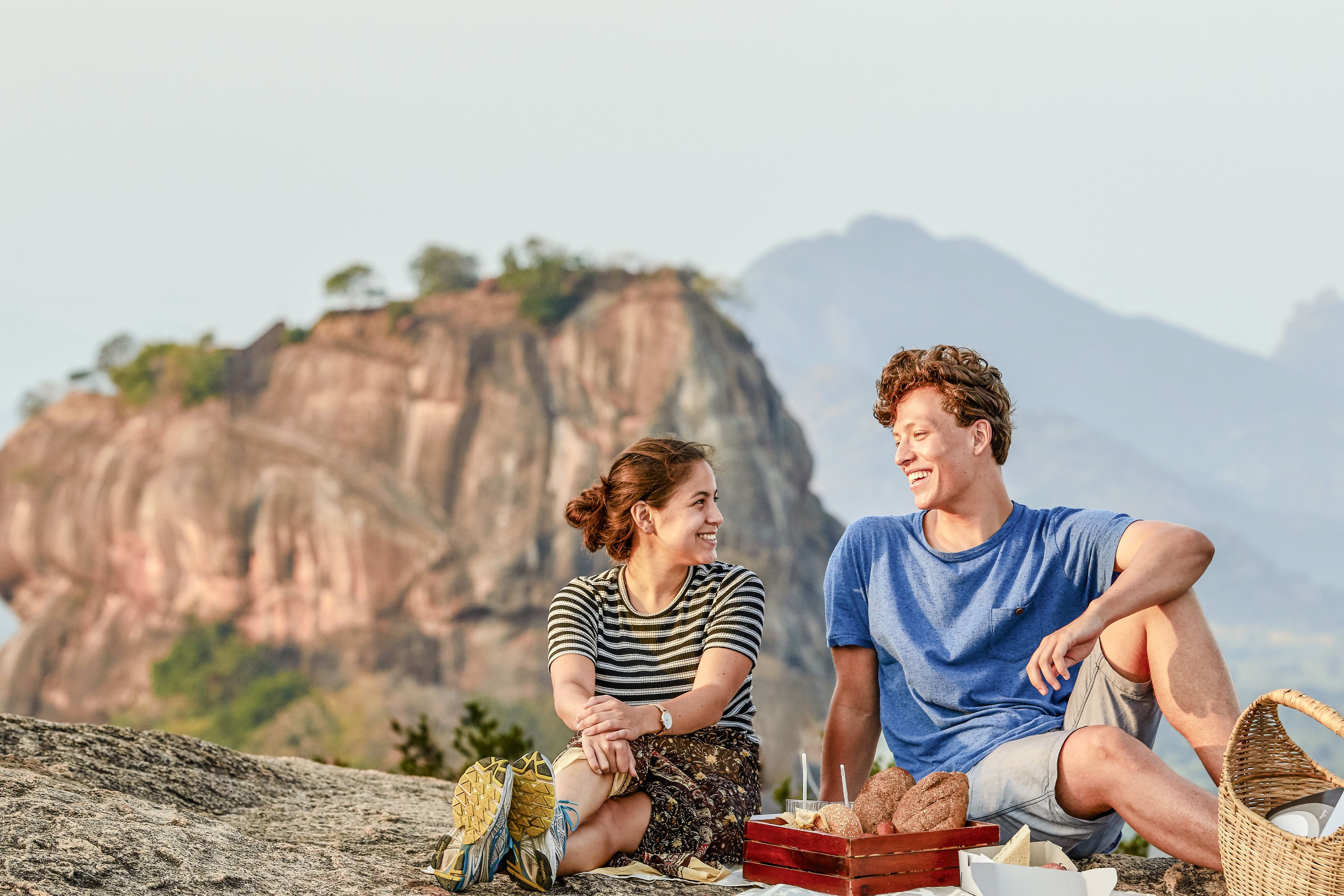 Two people sitting on a rocky ledge with a mountainous backdrop, enjoying a picnic with bread and drinks.