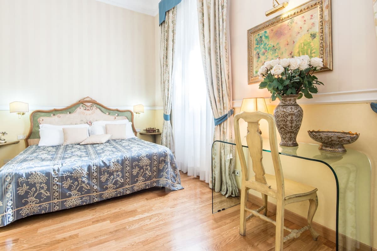 A hotel room with a bed, a chair, a table with a vase, and a painting on the wall.