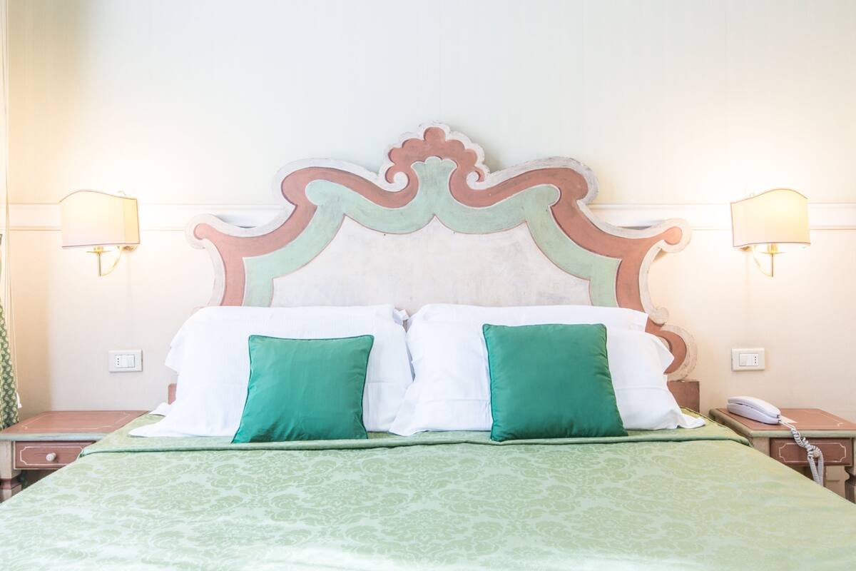 A hotel room with a bed, green and white bedding, a decorative headboard, bedside lamps, and a bedside telephone.