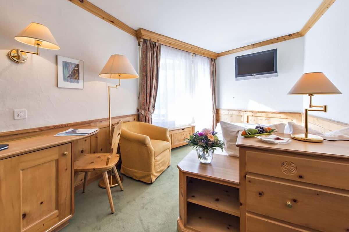 Een hotelkamer met een houten bureau, twee stoelen, een bed met een wit hoofdeinde, een flatscreen-tv en een raam met gordijnen.