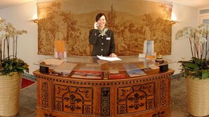 A reception desk with a person standing behind it, a large tapestry on the wall, and potted plants on either side. - Quadratscha Hotel (Samedan)