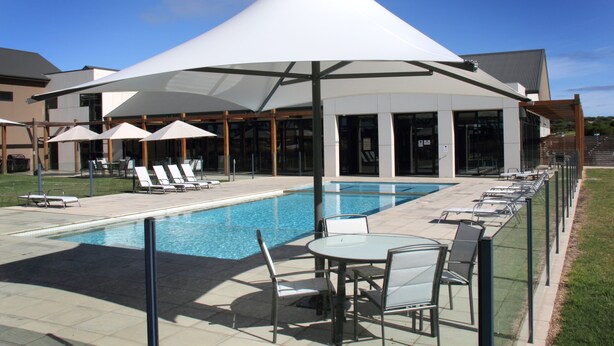 A pool area with lounge chairs and umbrellas, a covered seating area, and a modern building in the background. - Barwon Heads Resort at 13th Beach (Connewarre)