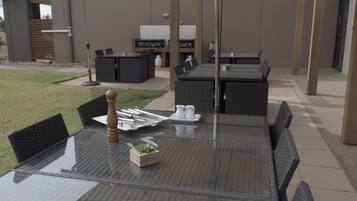 An outdoor patio with a glass table, wicker chairs, and a grill area in the background.