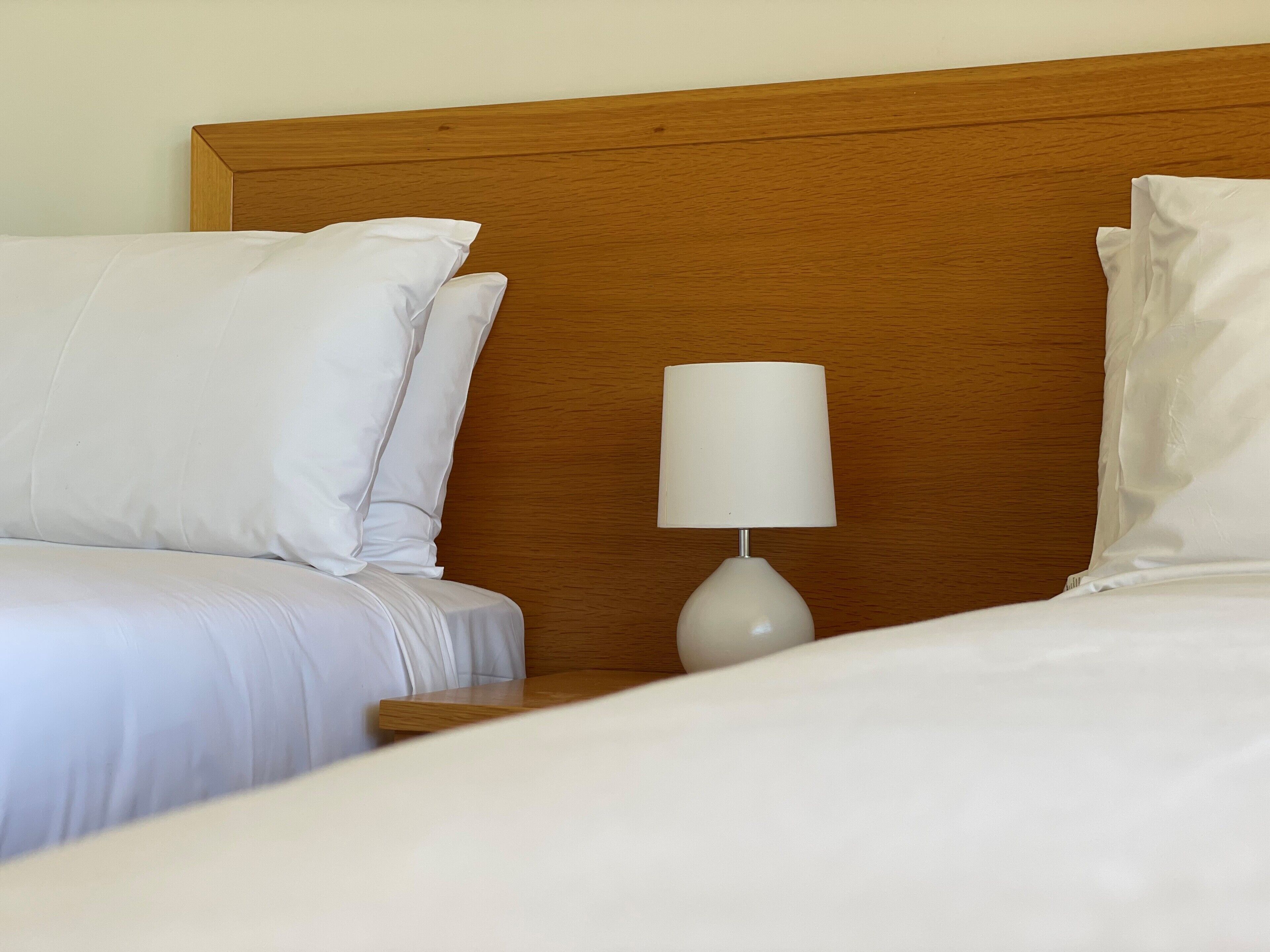 A neatly made twin bed with white linens, a wooden headboard, and a bedside table with a white lamp.