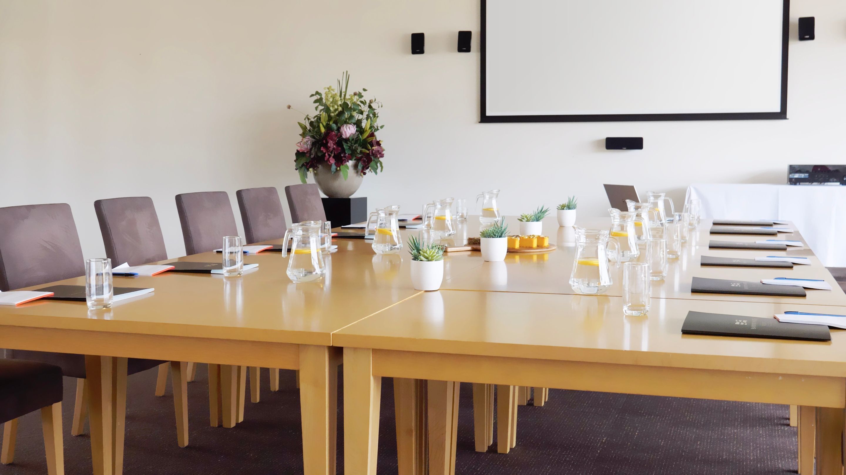Un salón de conferencias con una larga mesa, sillas, jarras de agua y una pantalla de proyector.