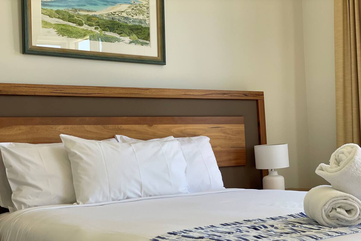 A neatly made bed with white linens and pillows, a wooden headboard, a framed landscape painting, a bedside lamp, and a rolled towel on the side.