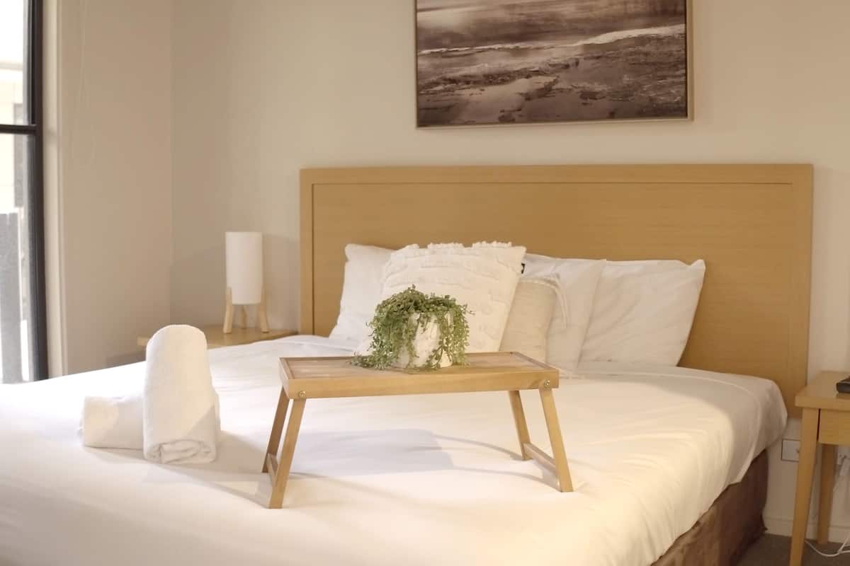A neatly made bed with white linens, a wooden bedside table with a plant, and a painting on the wall.