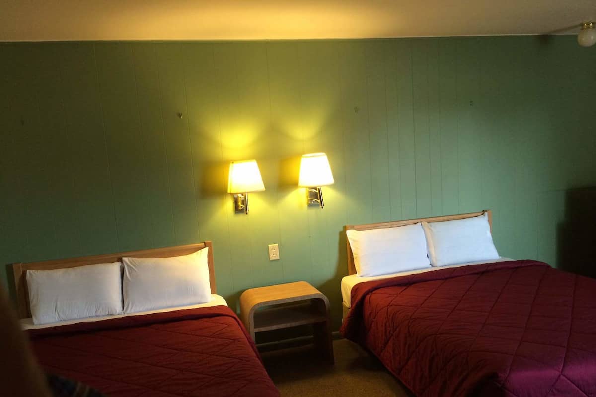 A hotel room with two beds, each with a red quilted bedspread and white pillows. There are two wall-mounted lamps on either side of the beds.
