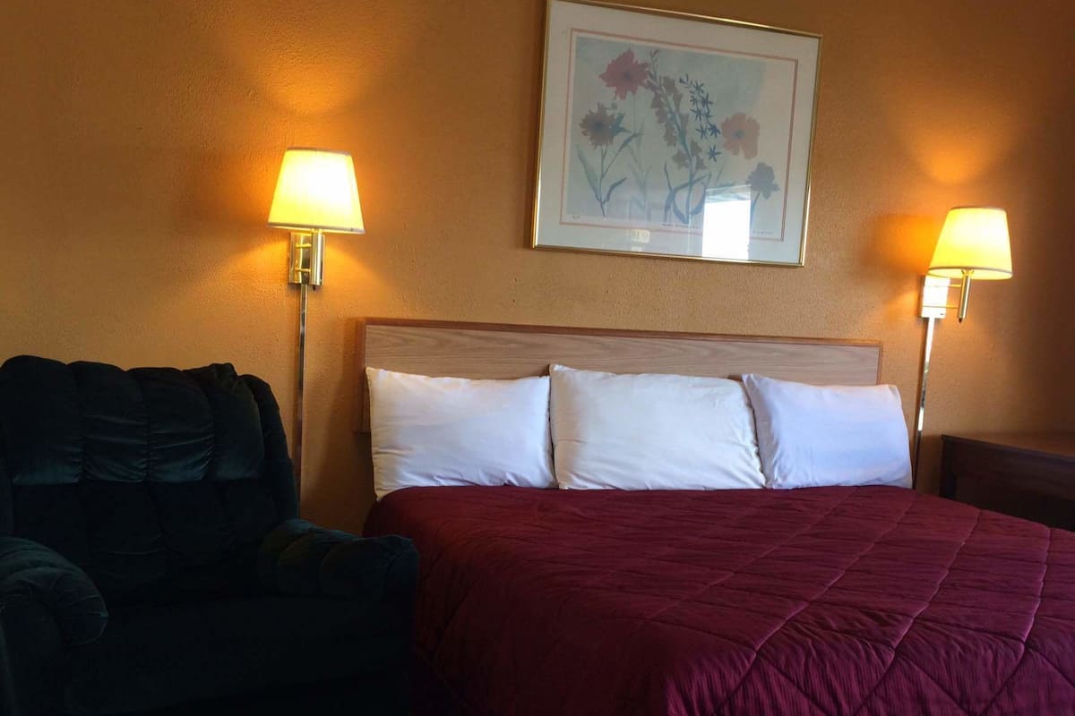A hotel room with a large bed, two bedside lamps, a framed floral artwork on the wall, and a dark-colored armchair.