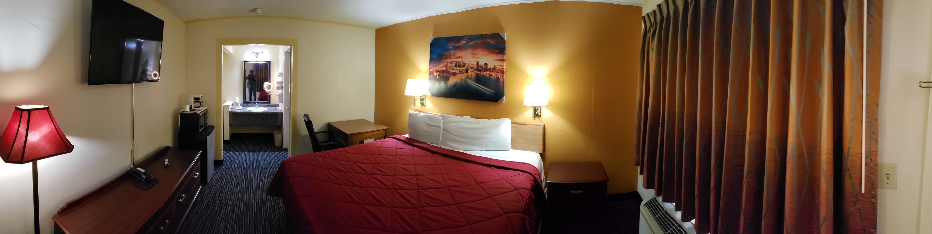 Une chambre d’hôtel avec un lit, un bureau, une chaise, une télévision, une salle de bain avec un miroir et un tableau au mur.