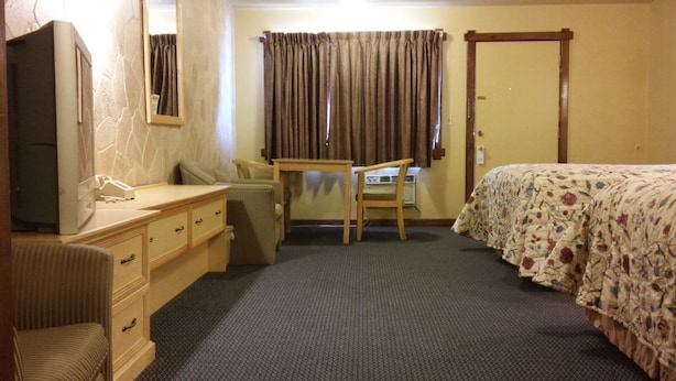 A hotel room with a bed, a desk, a chair, a dresser, and a television. - Budget Host Longhorn Motel (Byers)
