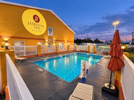 A swimming pool area with lounge chairs and umbrellas, surrounded by a white fence, and a building with a yellow exterior and a sign reading 'Jameson Inn & Suites'.