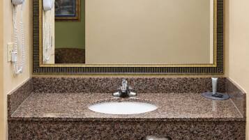 A bathroom with a granite countertop sink, a mirror with a gold frame, a hairdryer, and a trash can.