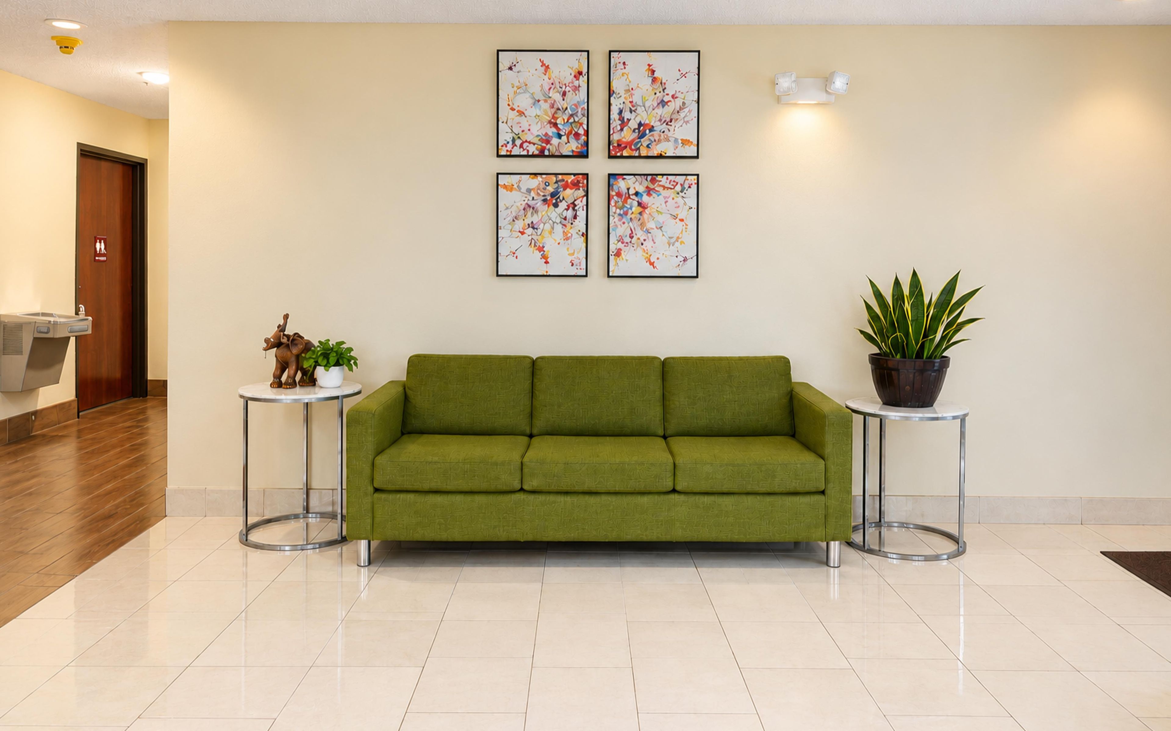 A green sofa in a lobby with a tiled floor, a small table with a potted plant, and abstract art on the wall.