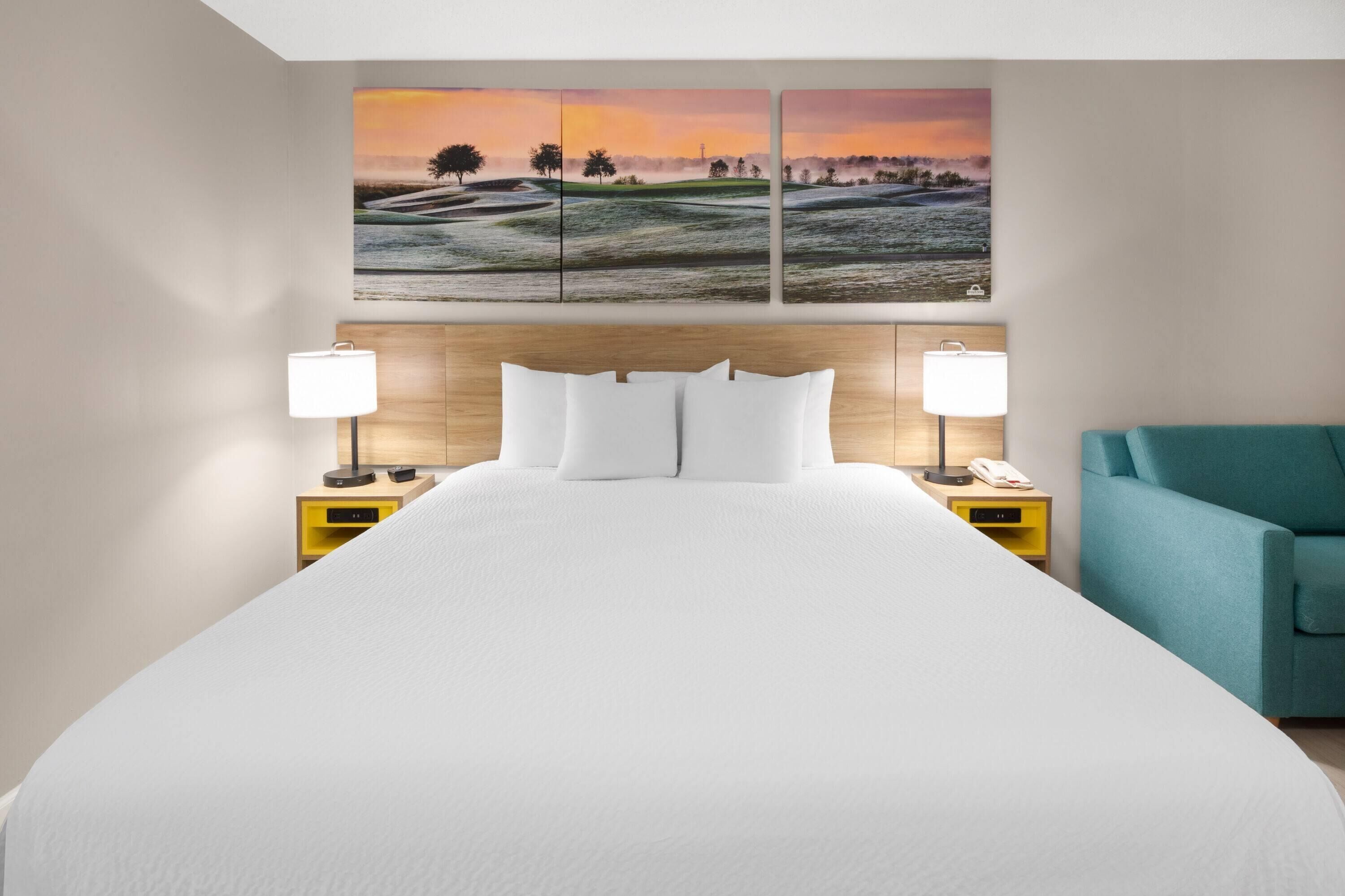 Una cama grande con ropa blanca, dos mesitas de noche con lámparas, un sillón color turquesa y un cuadro de un paisaje sobre la cama.