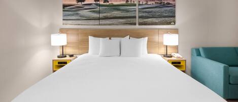 Una cama grande con ropa blanca, dos mesitas de noche con lámparas, un sillón color turquesa y un cuadro de un paisaje sobre la cama.