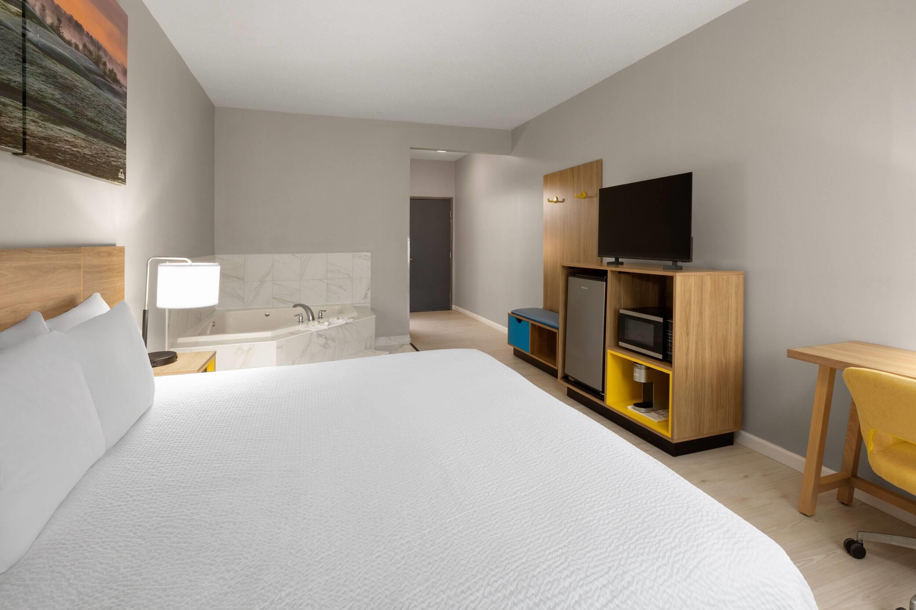 A modern hotel room with a large bed, a built-in desk, a flat-screen TV, a minibar, and a bathroom with a bathtub and shower.