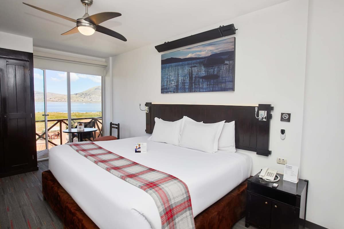 A hotel room with a large bed, a ceiling fan, a painting, a nightstand with a phone, and a view of the outdoors.