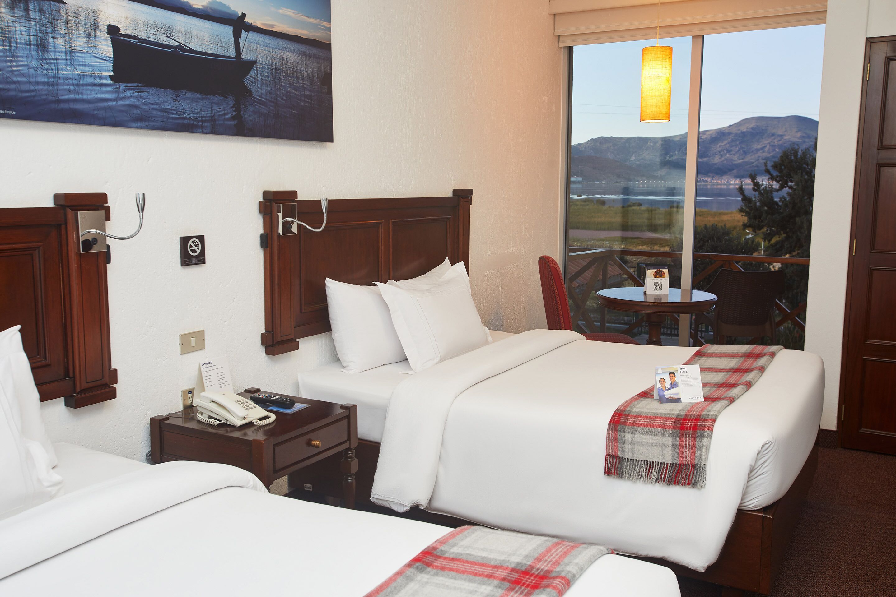 Habitación de hotel con dos camas, una mesita, una silla, un cuadro de un barco y vistas a montañas y una masa de agua.