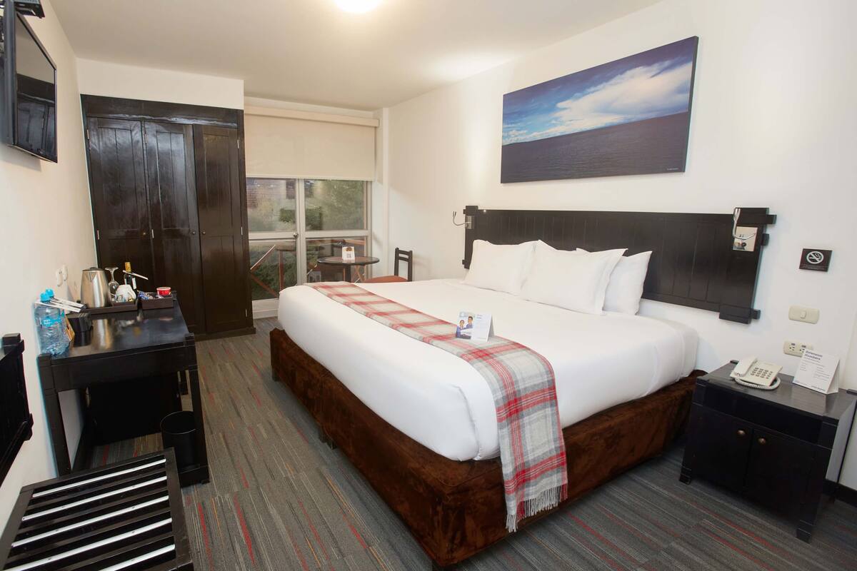 A hotel room with a large bed, a desk, a chair, a small table, a nightstand, and a window with a view of the outdoors.