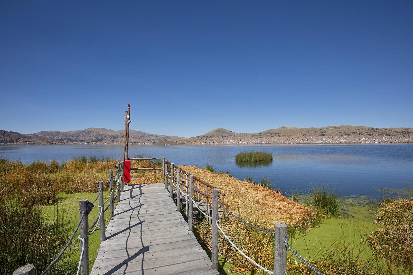 A wooden boardwalk leading to a lake with a clear view of distant hills and a small island.