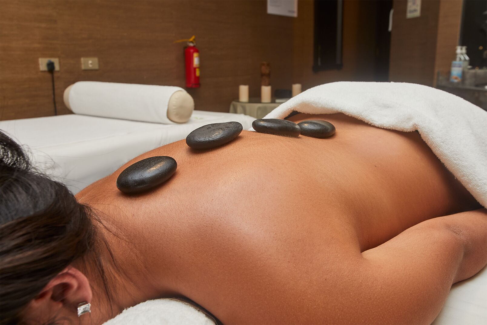 A massage therapy session with stones on a client's back, a fire extinguisher, and a white towel on the bed.