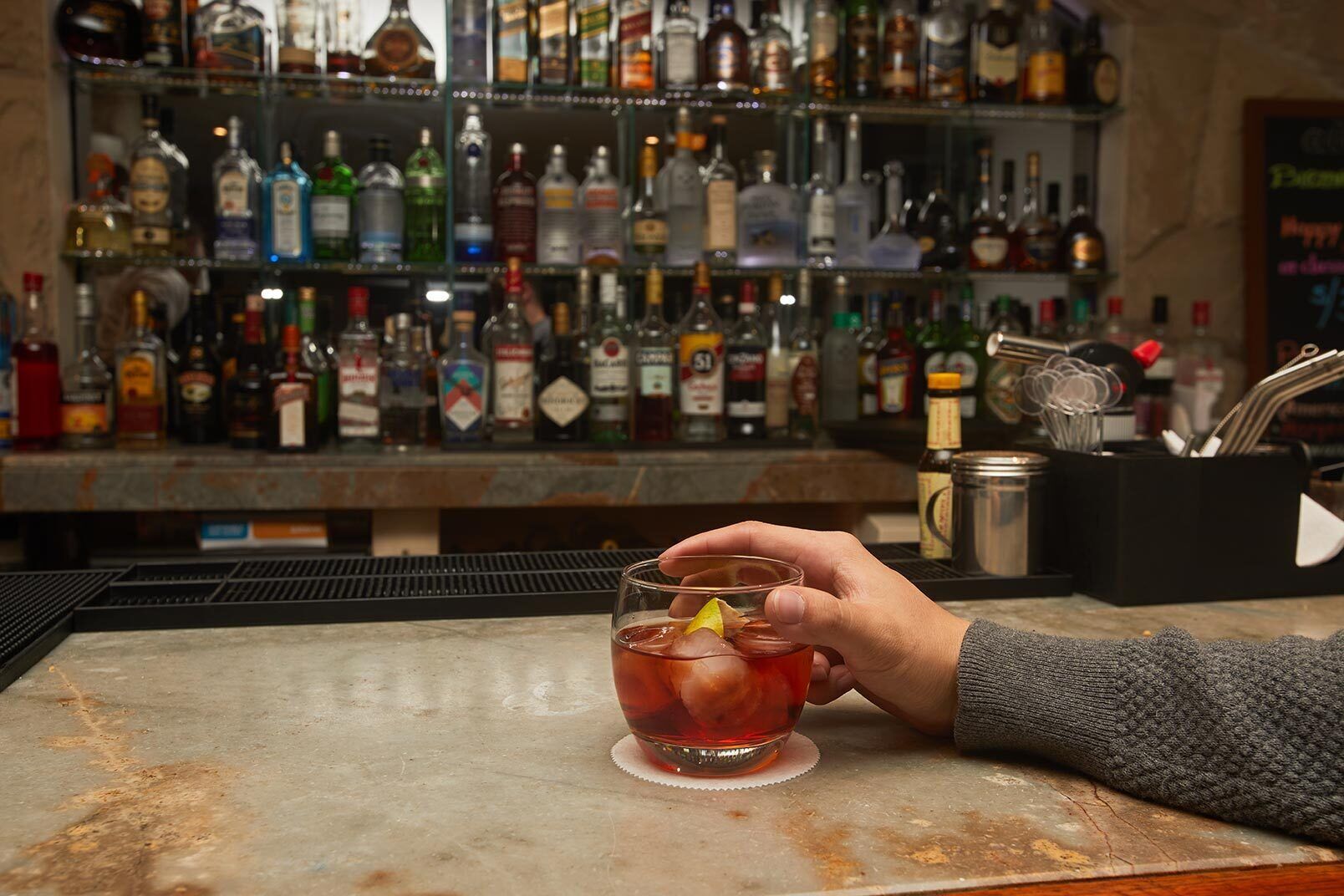 A bar area with a hand holding a drink, a variety of liquor bottles in the background, and bar tools on the counter.