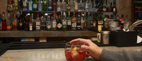A bar area with a hand holding a drink, a variety of liquor bottles in the background, and bar tools on the counter.