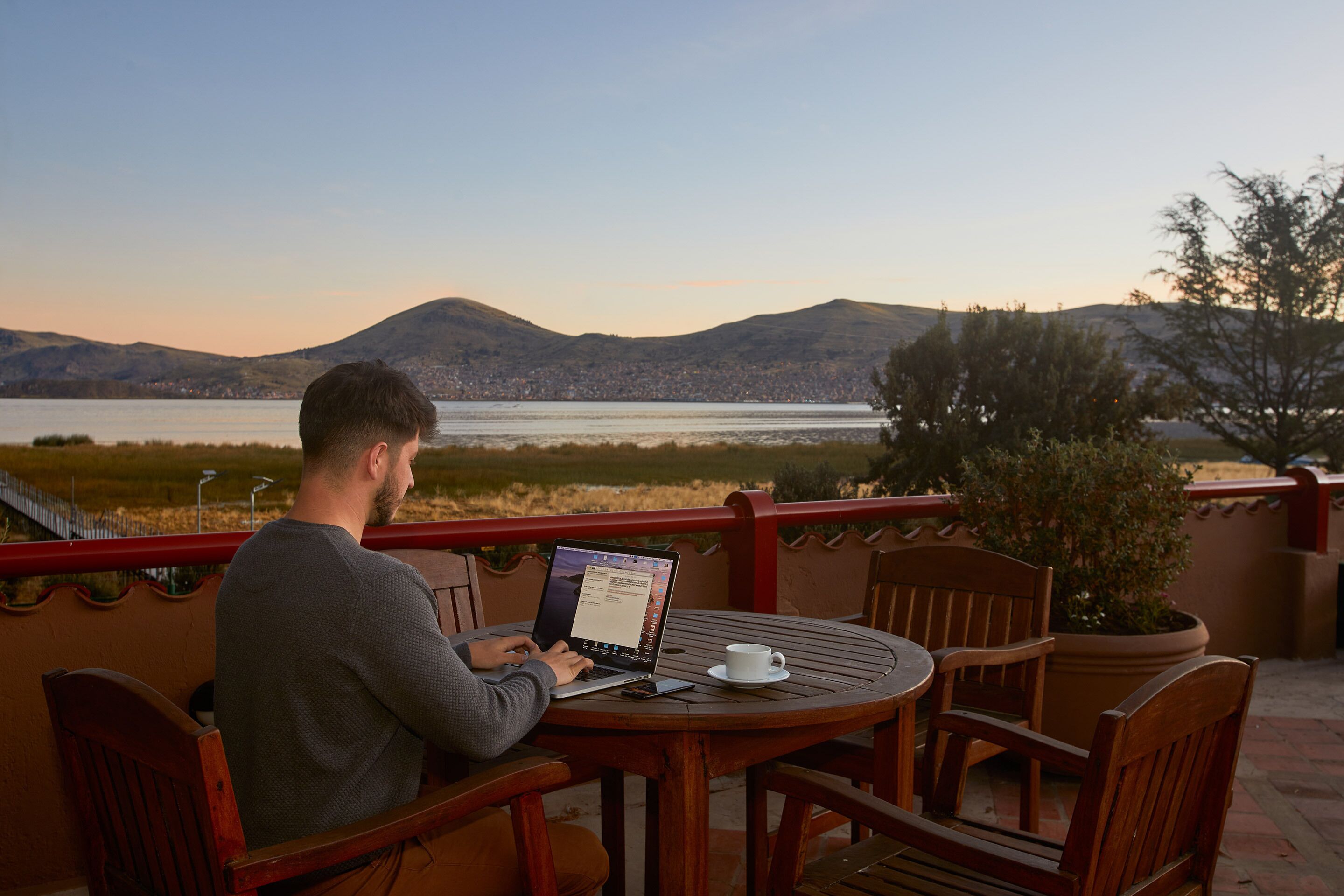 A man using a laptop on a wooden table with a white cup and saucer, overlooking a lake with mountains in the background.