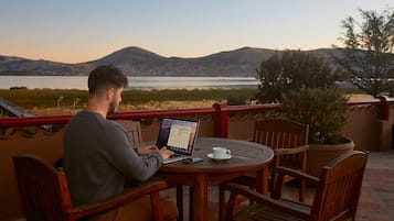 A man using a laptop on a wooden table with a white cup and saucer, overlooking a lake with mountains in the background.