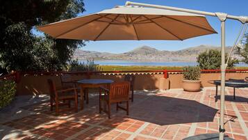 Un patio con mesa y sillas, una sombrilla grande y vistas a un lago y montañas.