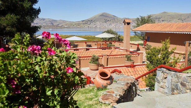 A courtyard with terracotta-roofed buildings, a stone staircase, and a view of a lake and hills. - Casa Andina Premium Puno (Puno)