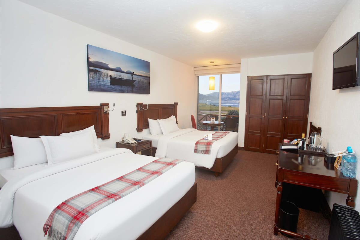 A hotel room with two beds, a desk, a TV, and a painting of a lake.