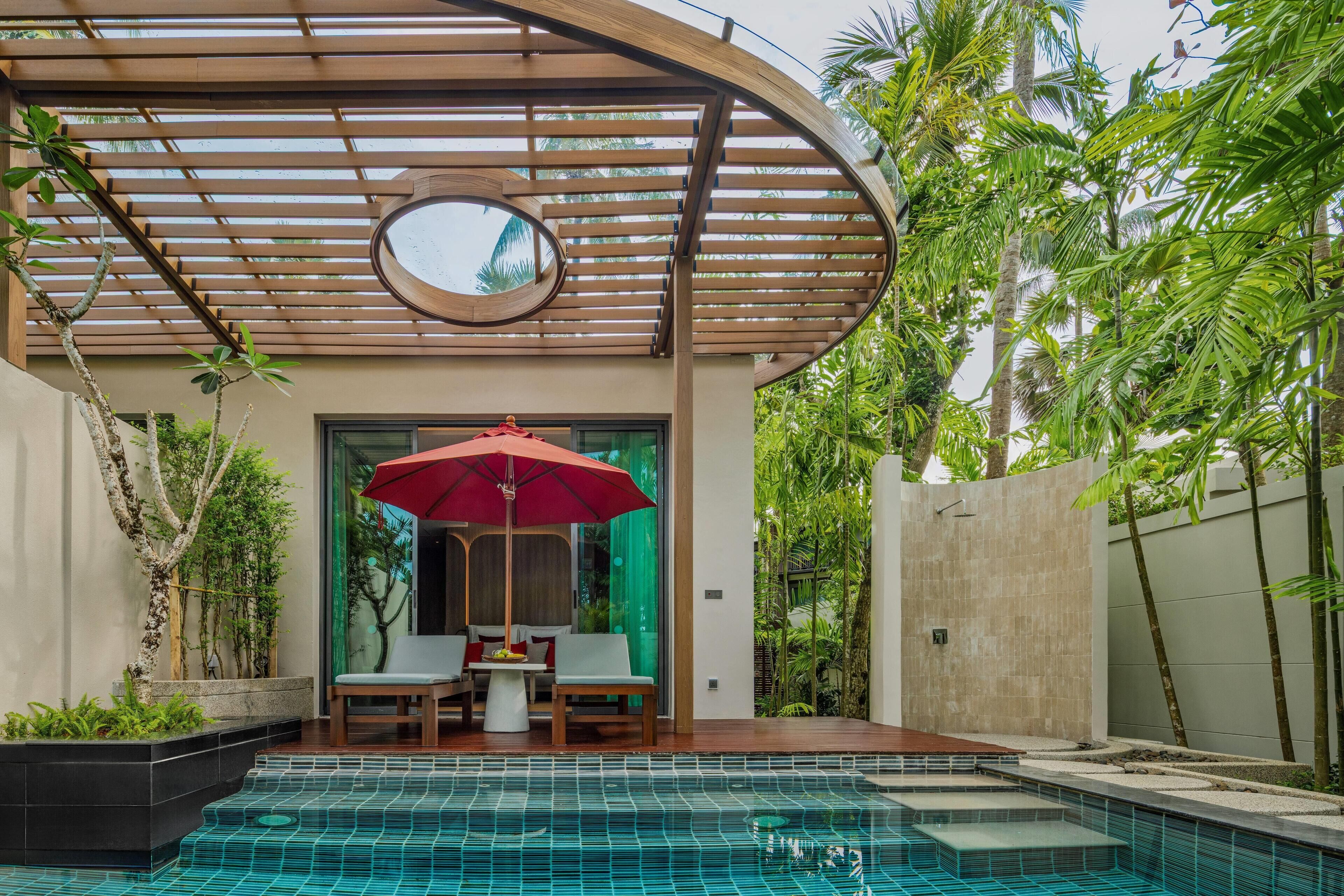 Un área de piscina moderna al aire libre con una pérgola de madera, una sombrilla roja y un espacio de estar con dos sillas y una mesa.