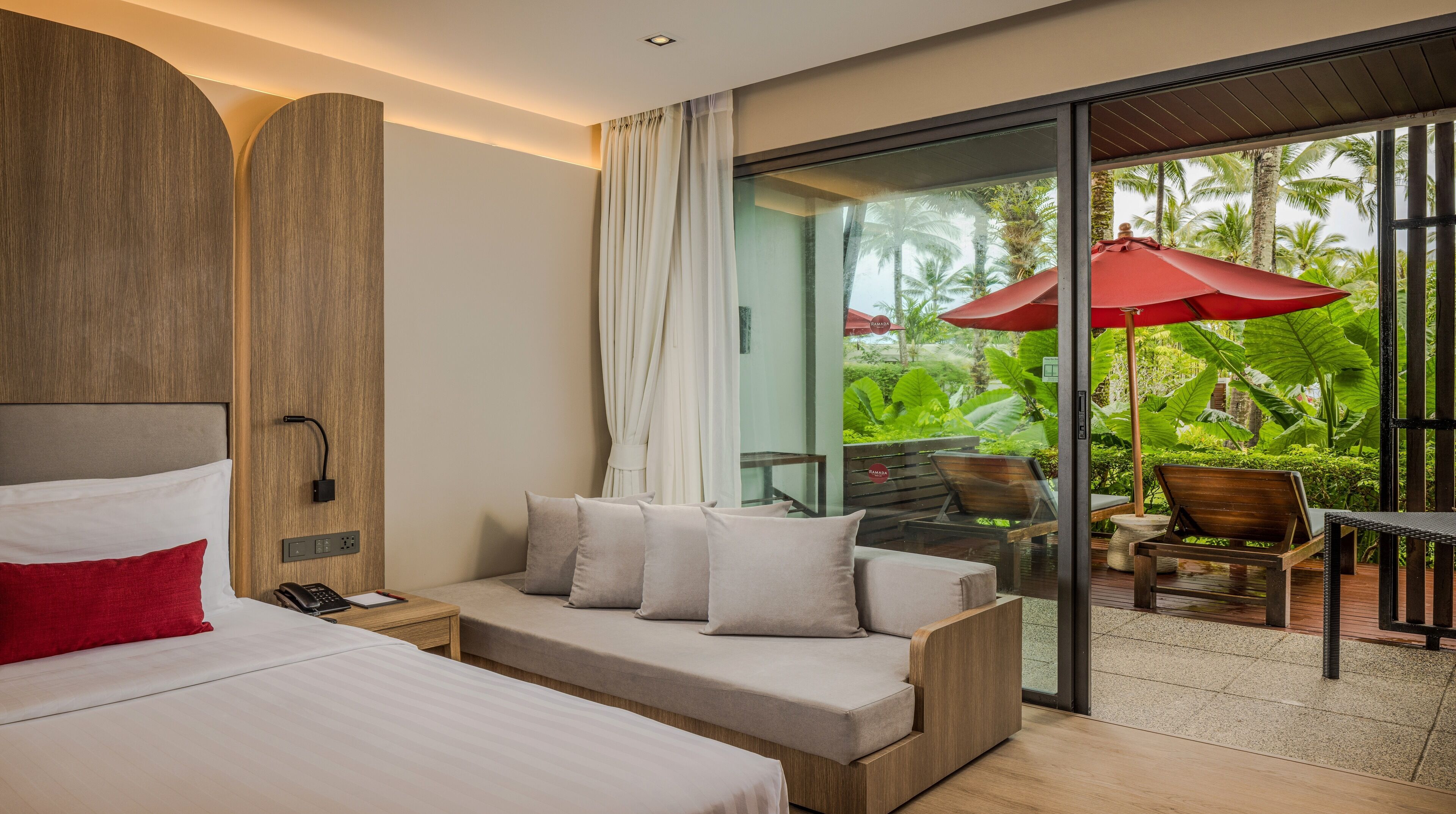 A modern hotel room with a bed, a sofa, a glass door leading to a balcony with outdoor furniture and a red umbrella.
