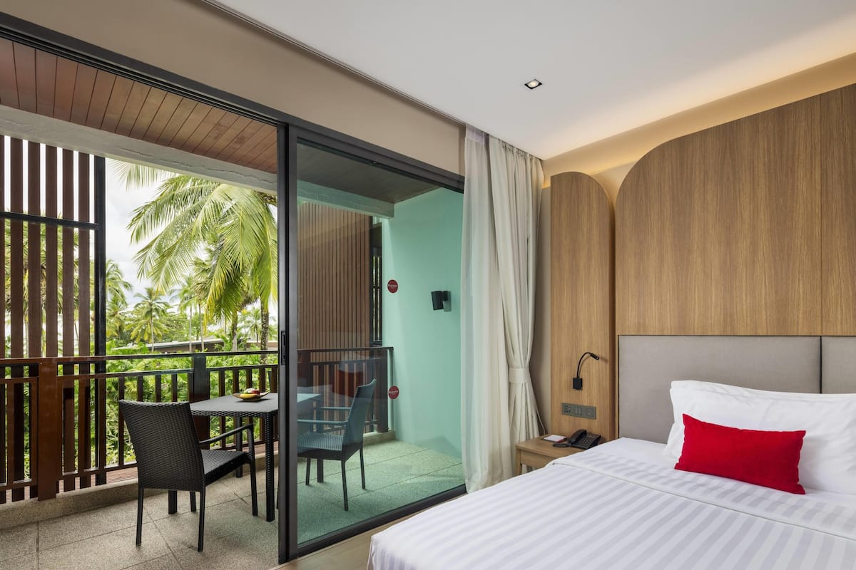 A hotel room with a bed, a desk with a chair, a balcony with a table and chairs, and a view of palm trees.