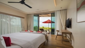 Ein Hotelzimmer mit einem großen Bett, einem Schreibtisch, einem Sessel und einem Fernseher. Ein Balkon mit einem roten Sonnenschirm bietet Blick auf den Ozean.