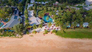Eine Vogelperspektive zeigt einen luxuriösen Resortkomplex mit einem breiten Sandstrand, einem von Palmen umgebenen Pool und mehreren Villen direkt am Strand.