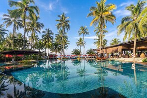 Une piscine entourée de palmiers et un bâtiment d’hôtel doté d’un espace extérieur aménagé avec des sièges.