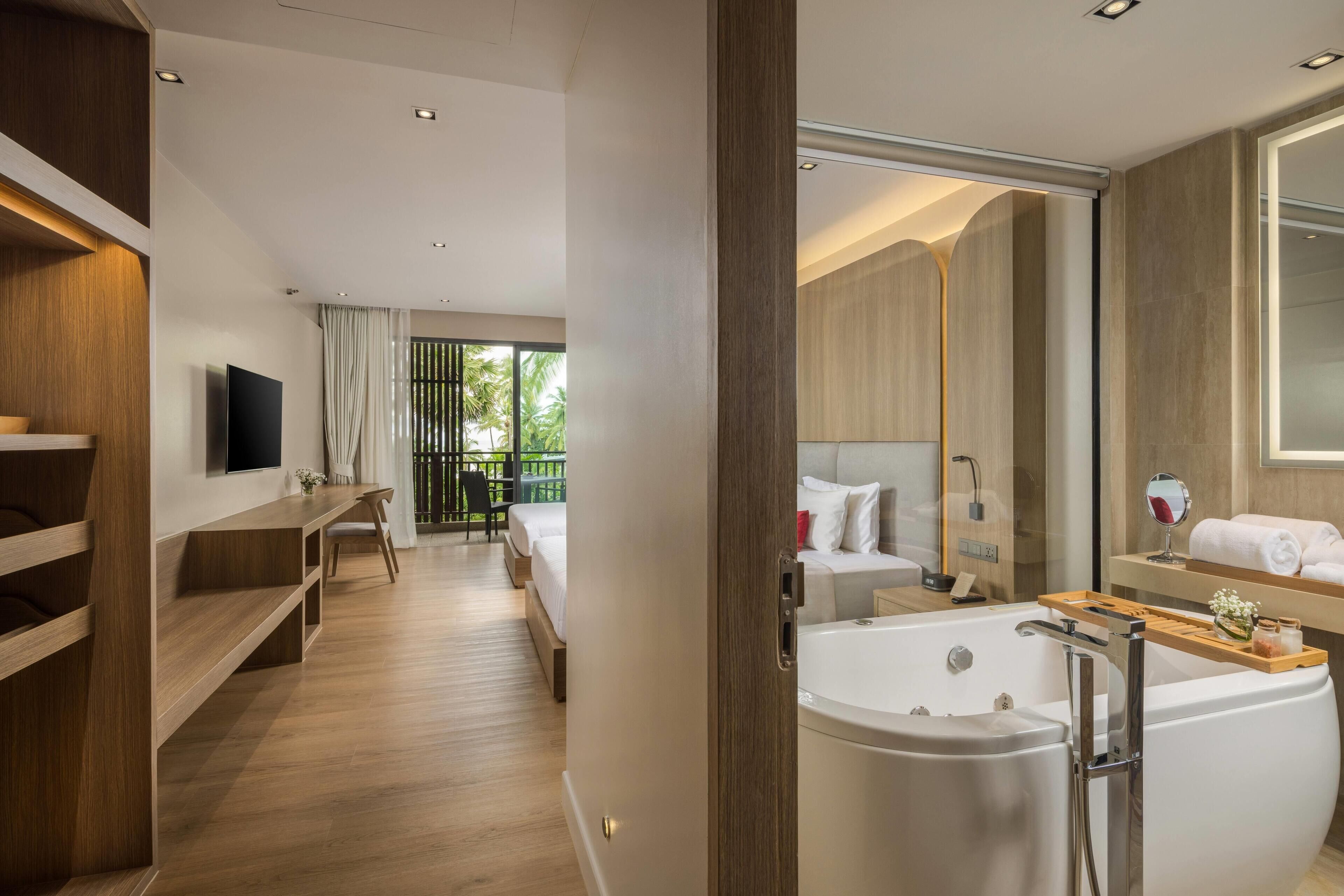 Una habitación de hotel moderna con suelo de madera, una bañera grande, un televisor de pantalla plana, un balcón con vistas a palmeras y un banco de madera.