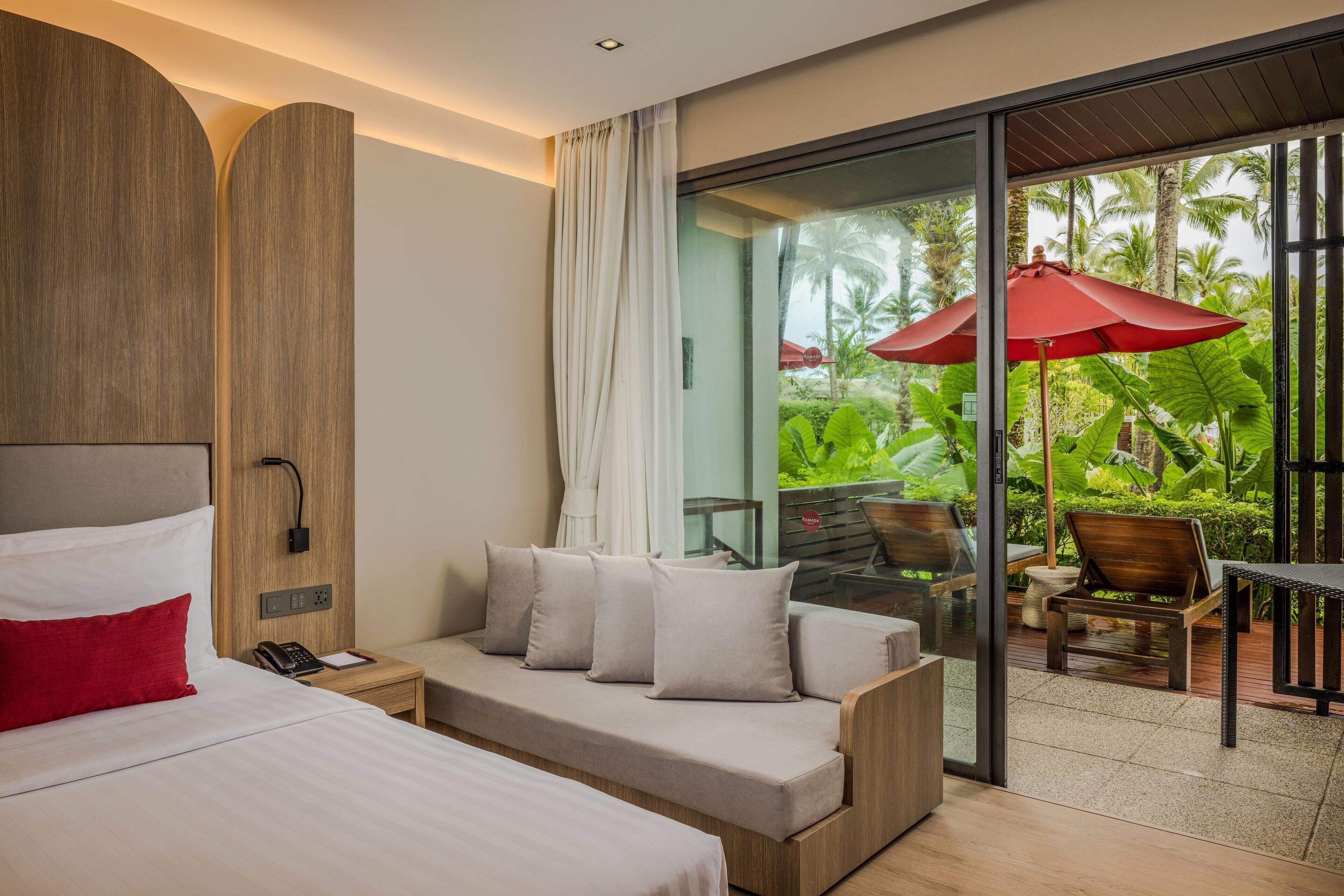 Una habitación de hotel moderna con una cama, un sofá, una mesita y un vidrio que da a un balcón con mobiliario exterior y vegetación.