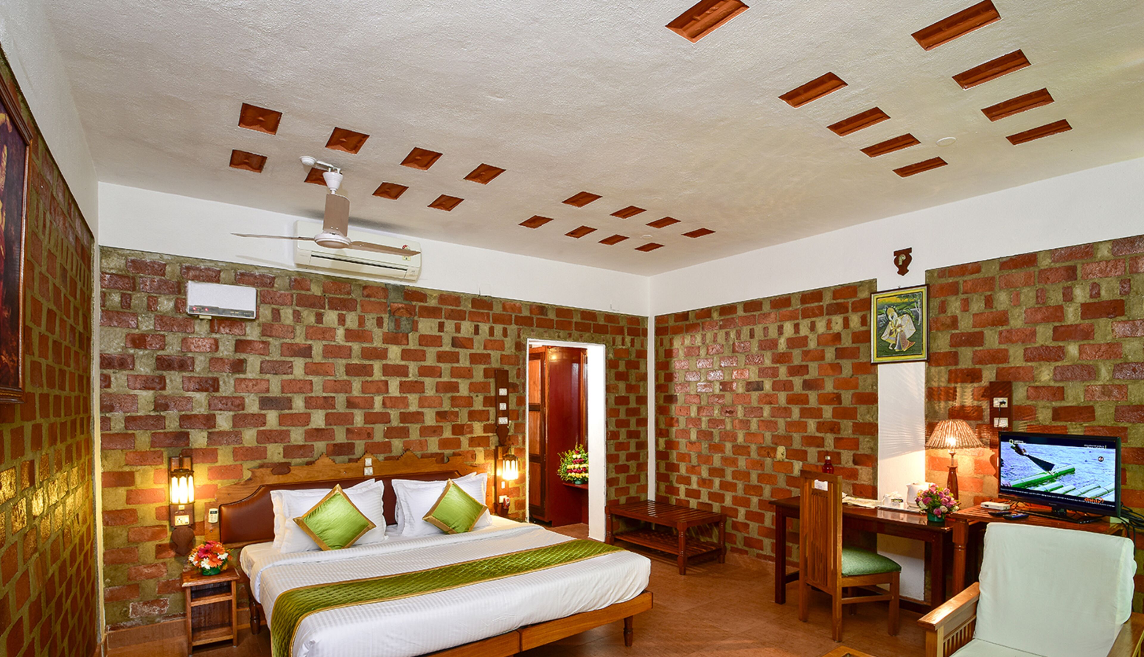 A hotel room with a brick-patterned wall, a bed with green pillows, a desk with a computer, and a TV.