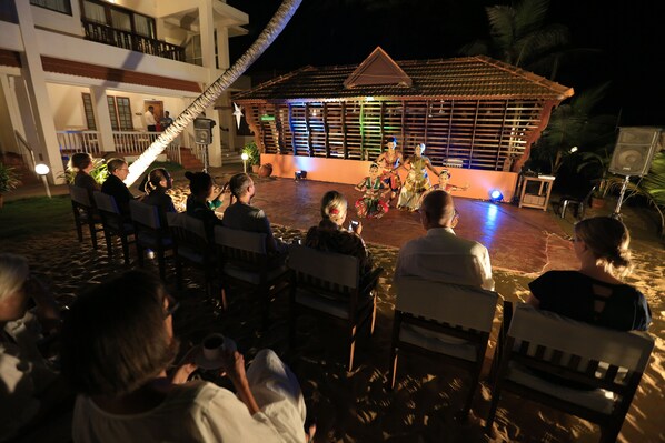 A stage with performers in traditional attire, an audience seated on chairs, and a building with a tiled roof in the background. - Beach and Lake Ayurvedic Resort (Thiruvananthapuram)