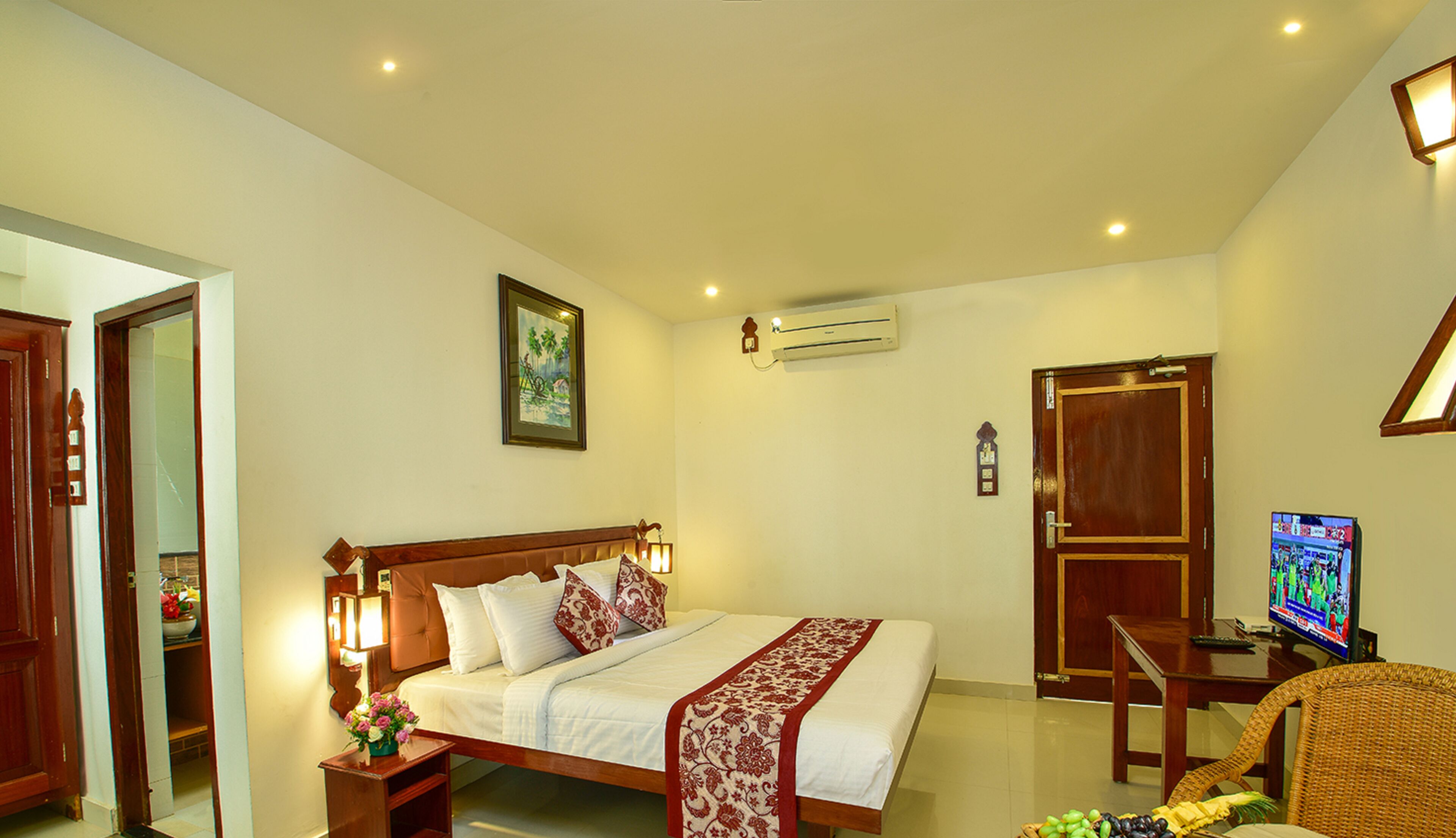 A hotel room with a bed, bedside table, lamp, air conditioning unit, TV, and a chair.