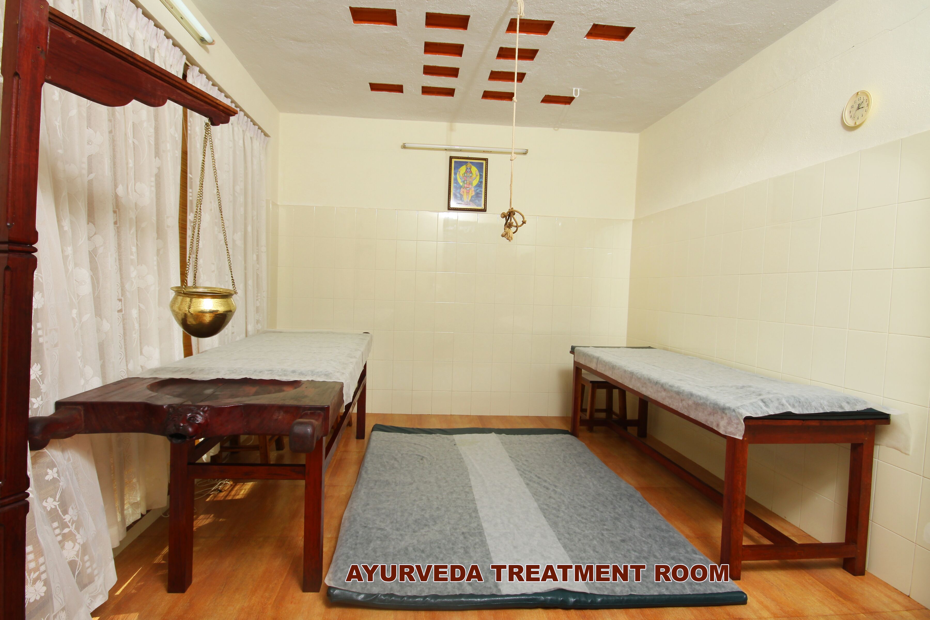 Une salle de soins ayurvédique équipée de deux lits en bois, d’un pot en laiton suspendu et d’un tableau encadré au mur.