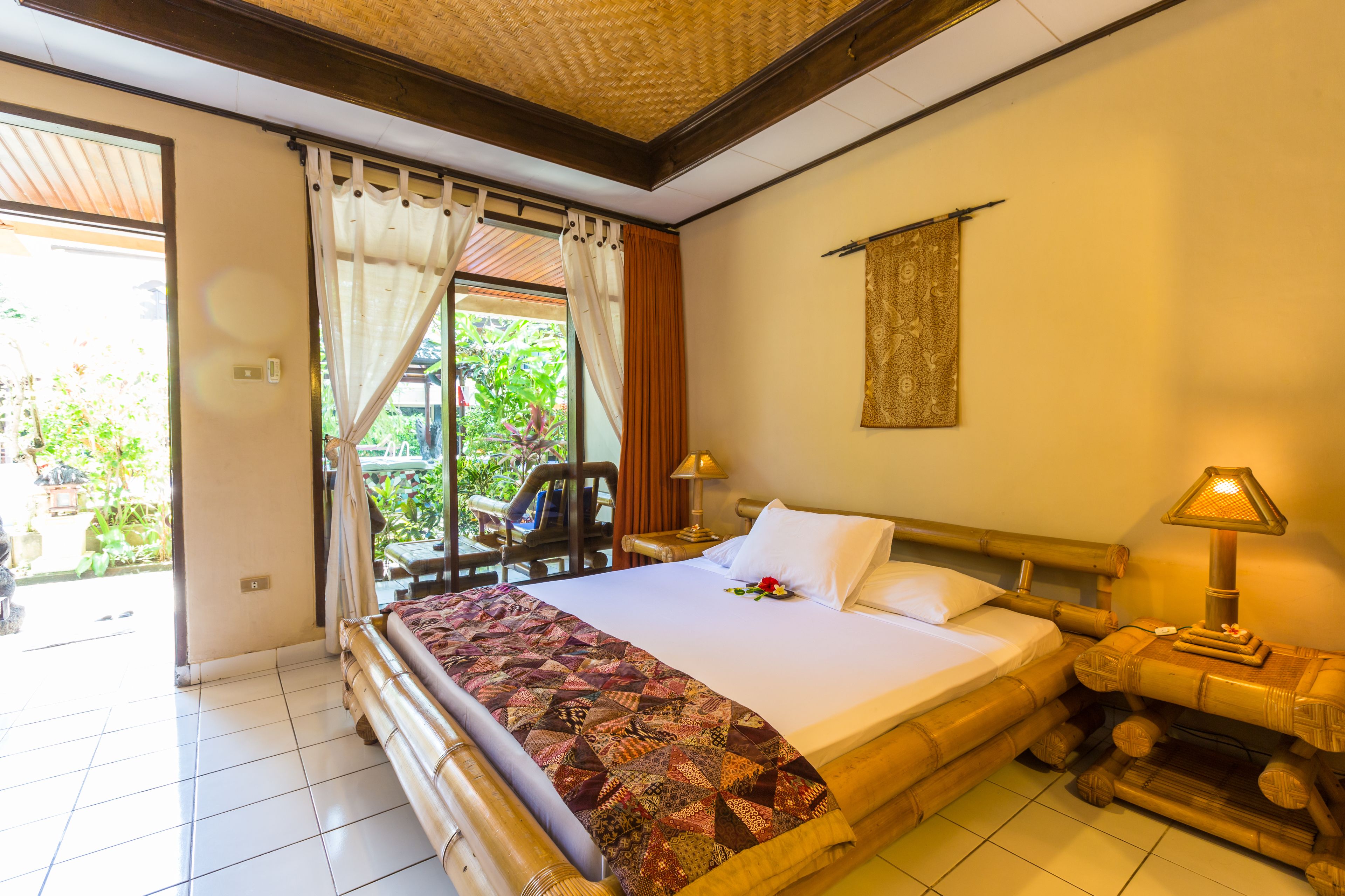 A hotel room with a large bed, a wooden headboard, a nightstand with a lamp, and a view of greenery outside.