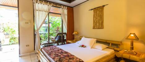 A hotel room with a large bed, a wooden headboard, a nightstand with a lamp, and a view of greenery outside.